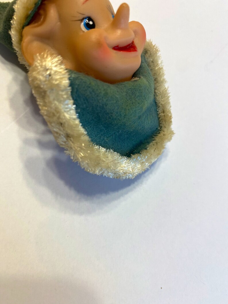 May include: A vintage elf ornament with a green felt hat and a white fur trim. The elf has a large nose, blue eyes, and a rosy-cheeked smile.