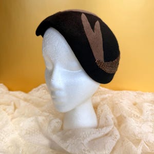 May include: A black beret-style hat with a brown accent on top and two brown decorative shapes. The hat features a beaded embellishment along the bottom edge. The hat is displayed on a white mannequin head.