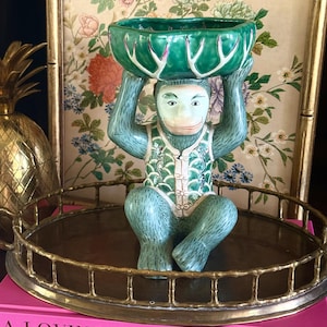 May include: A ceramic monkey figurine in a seated position, holding a green bowl above its head. The monkey is light blue with a detailed green vest. The bowl has a white and green rim. The figurine sits on a gold-colored tray.