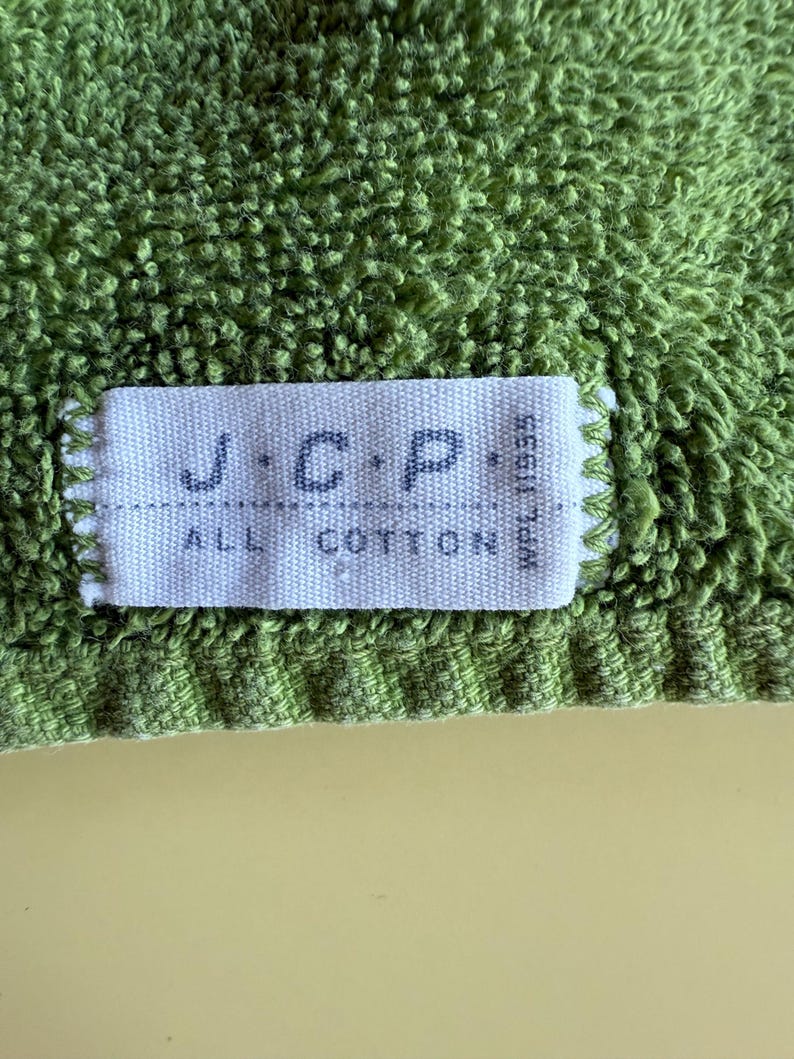 May include: Close-up of a green terry cloth towel with a white label. The label reads "J.C.P. ALL COTTON" in dark blue letters. The towel has a textured surface and a light green color.