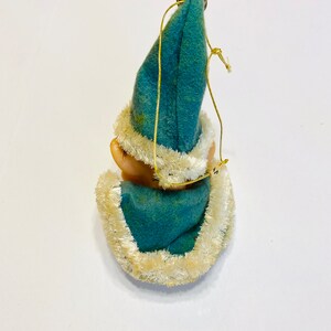 May include: A blue felt Christmas ornament with a white fur trim, depicting a small elf wearing a blue hat. The ornament has a gold string for hanging.