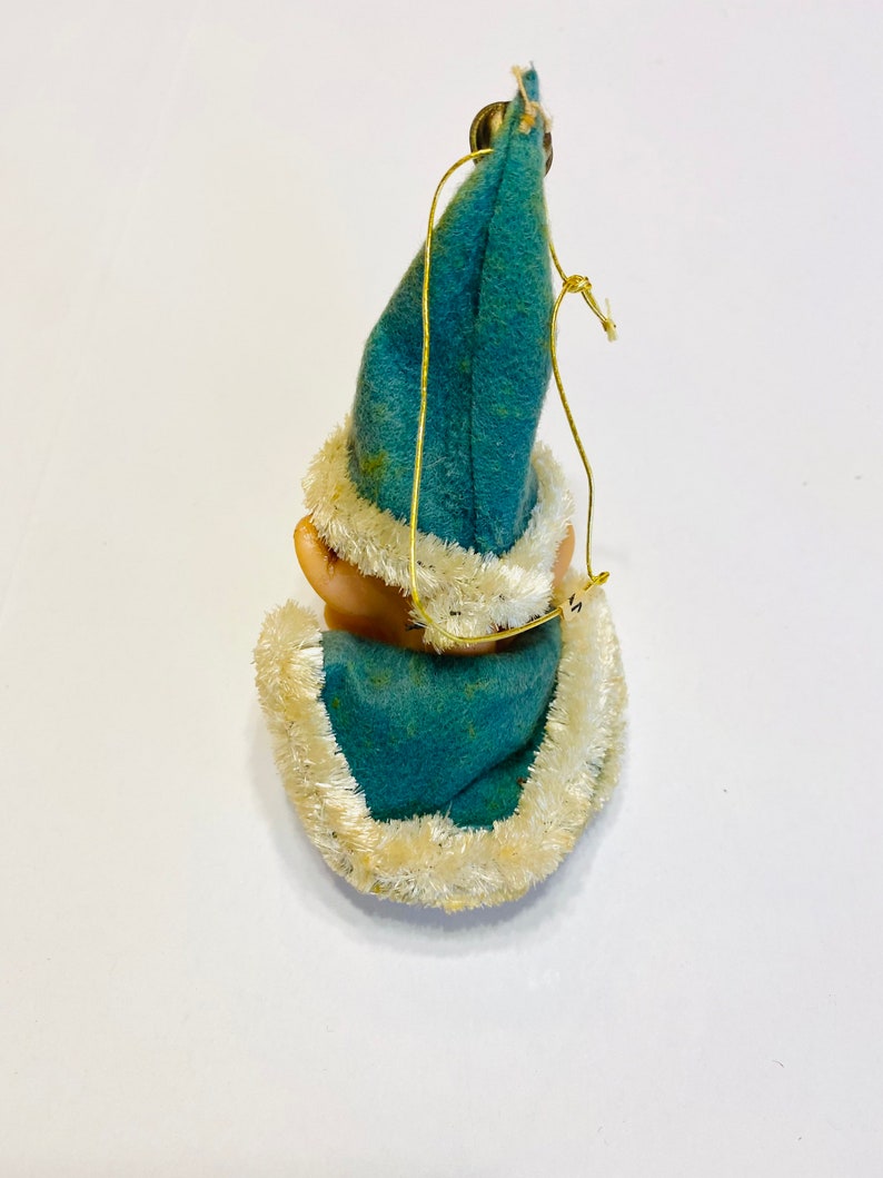 May include: A blue felt Christmas ornament with a white fur trim, depicting a small elf wearing a blue hat. The ornament has a gold string for hanging.