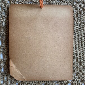 May include: A rectangular, light brown card with a small, orange loop at the top. The card has a slightly textured surface and a torn corner. The background is a beige, crocheted doily.