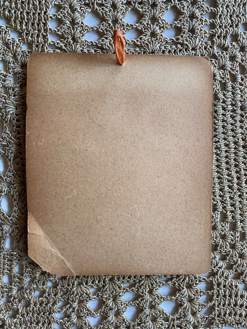 May include: A rectangular, light brown card with a small, orange loop at the top. The card has a slightly textured surface and a torn corner. The background is a beige, crocheted doily.