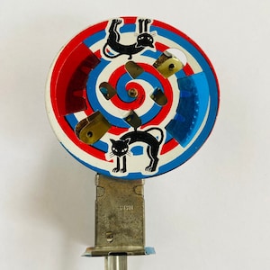 May include: A vintage tin toy with a red, white, and blue spinning wheel featuring black cats. The wheel is attached to a metal base with a key wind-up mechanism.