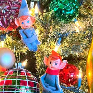 May include: A decorated Christmas tree with various ornaments. The ornaments include vintage elf figurines in blue felt outfits with red hair and hats, and colorful glass ball ornaments in red, green, and gold. The tree is lit with small white lights.