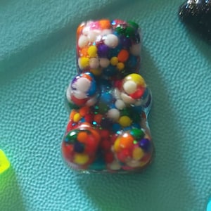 May include: A colorful gummy bear charm with red, yellow, blue, green, and white sprinkles. The charm is made of clear resin and has a glossy finish.