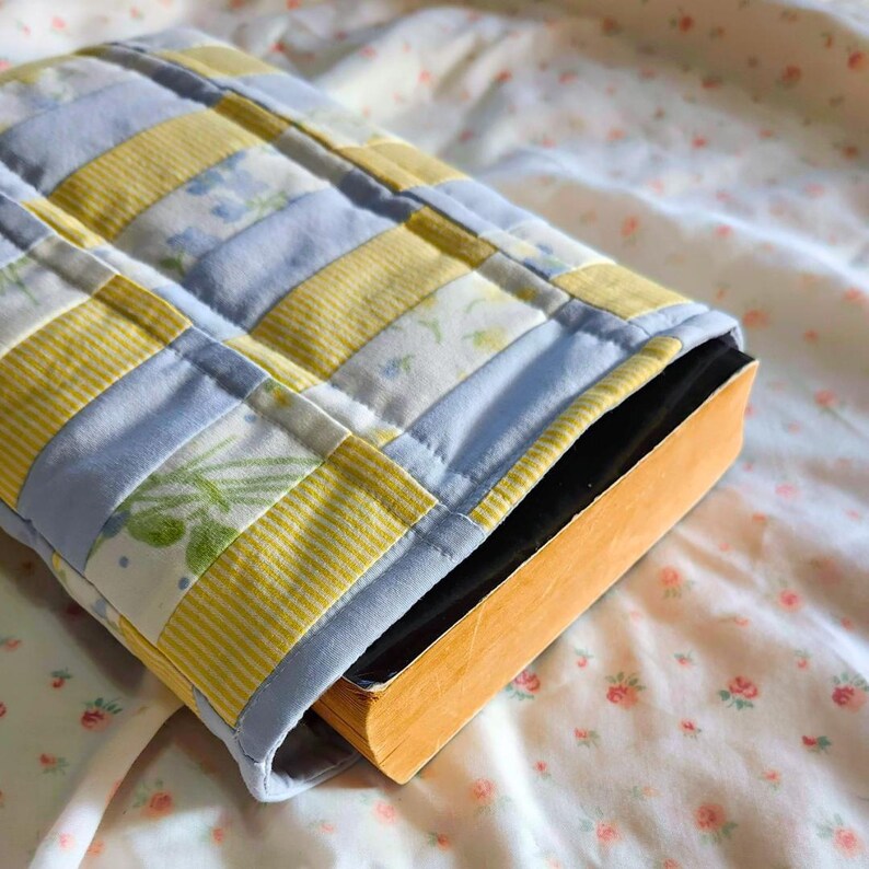 Peut inclure: Une housse de livre en tissu matelass&eacute; pr&eacute;sentant un motif patchwork compos&eacute; de bandes altern&eacute;es jaunes et bleu clair. Les sections jaunes incluent des motifs unis et ray&eacute;s, tandis que les sections bleu clair pr&eacute;sentent un imprim&eacute; floral discret. Un livre est ins&eacute;r&eacute; &agrave; l'int&eacute;rieur.