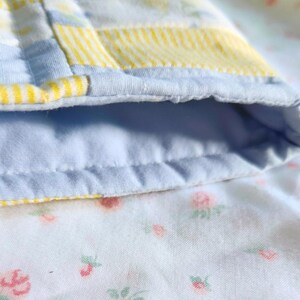 Peut inclure: Une pi&egrave;ce de tissu matelass&eacute;e pr&eacute;sentant un motif patchwork avec des sections ray&eacute;es bleu clair et jaune. Le bord est fini avec un biais bleu clair uni. L'envers du tissu affiche un fond blanc avec un petit motif floral imprim&eacute;.
