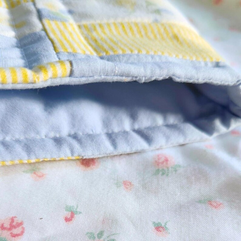 Peut inclure: Une pi&egrave;ce de tissu matelass&eacute;e pr&eacute;sentant un motif patchwork avec des sections ray&eacute;es bleu clair et jaune. Le bord est fini avec un biais bleu clair uni. L'envers du tissu affiche un fond blanc avec un petit motif floral imprim&eacute;.