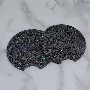 May include: Two black glitter car coasters with a cut-out for a cup holder. The coasters have a sparkly, iridescent finish.
