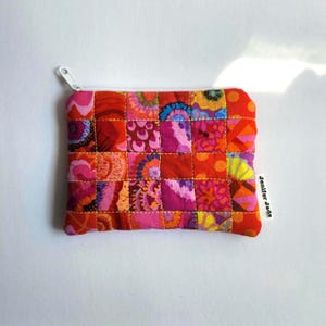 May include: A rectangular fabric pouch featuring a quilted patchwork design with various floral and abstract patterns in shades of red, orange, pink, and yellow. The pouch includes a white zipper closure and a small white tag labeled Jenifer Jade.