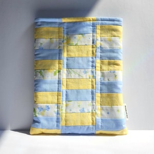 May include: A rectangular quilted fabric sleeve featuring a patchwork design of alternating horizontal strips in light blue, yellow pinstripes, and a white floral print with small blue and yellow flowers. A small white fabric tag reads Dream of Jeni.