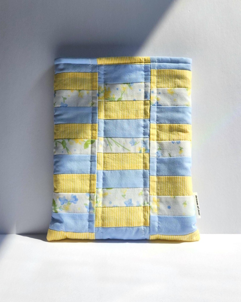 Peut inclure: Une pochette en tissu matelass&eacute; rectangulaire pr&eacute;sentant un motif patchwork de bandes horizontales altern&eacute;es bleu clair, &agrave; rayures jaunes et &agrave; imprim&eacute; floral blanc avec de petites fleurs bleues et jaunes. Une &eacute;tiquette Dream of Jeni est visible.