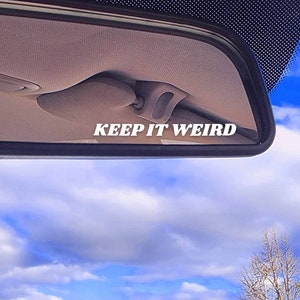 May include: A rearview mirror with a blue sky and white clouds reflected in it. The mirror has a sticker that says "KEEP IT WEIRD" in white letters.