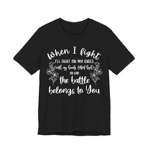 Op de afbeelding: Een zwart t-shirt met korte mouwen en witte tekst: When I fight I'll fight on my knees with my hands lifted high oh god the battle belongs to You. Het ontwerp wordt aan beide kanten geflankeerd door witte bloemillustraties.