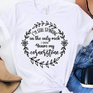 Pode incluir: Camiseta branca com um design de coroa de flores preta e o texto "I'm still standing on the only rock I know you're my cornerstone".