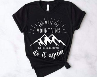 Può includere: Maglietta nera con testo bianco che recita "You move the mountains and I believe I'll see you do it again". Il testo è circondato da un design a raggiera stilizzato. La maglietta presenta anche una grafica di tre montagne bianche.