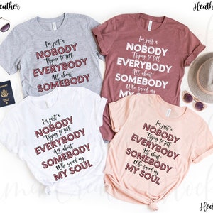 May include: Four t-shirts in different colors with the same text printed on them in a burgundy color. The text reads "I'm just a NOBODY Trying to tell EVERYBODY All about SOMEBODY Who saved my SOUL".