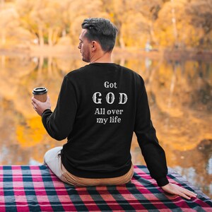 May include: A person wearing a black sweatshirt with the text "Got GOD All over my life" in white lettering. The person is sitting on a red and black plaid blanket in front of a lake.