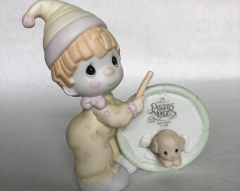 May include: A porcelain figurine of a clown with a yellow and white polka dot outfit holding a drum with a puppy peeking out of it. The drum has the text "The Enesco Precious Moments Birthday Club" on it.