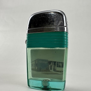 May include: A vintage lighter with a chrome top and a teal body. The lighter has a rectangular shape with a clear window showing a blurry image of a building. It has silver metal trim and a textured teal band around the middle.