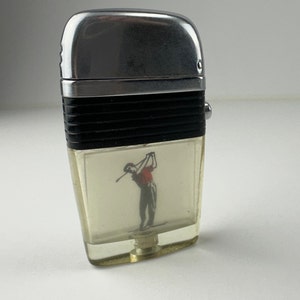 以下が含まれることがあります： 前面にゴルファーのグラフィックが描かれた銀と黒のライター。ライターには、ゴルファーが見える透明なプラスチック製の窓があります。