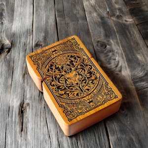 Puede incluir: Una caja rectangular de madera con tapa con bisagras. La tapa está intrincadamente tallada con un diseño central de toro rodeado de patrones florales y ornamentales. La caja es de madera clara y está sobre una superficie de madera envejecida.