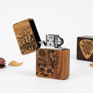 Puede incluir: Un encendedor de madera con un diseño de guitarra y flores. El encendedor es de madera marrón claro y tiene una parte superior de metal plateado. La tapa está abierta, revelando el interior. Un soporte de púas de guitarra de madera está en el fondo.