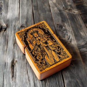 Puede incluir: Una caja de madera con tapa con bisagras, con un diseño grabado detallado. El diseño representa una figura vestida con una túnica que sostiene una espada, enmarcada por bordes ornamentados y elementos celestiales. La caja es de madera clara y descansa sobre una superficie de madera envejecida.