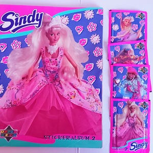 Könnte beinhalten: Ein rosa Sindy-Sticker-Album mit einer Puppenillustration und passenden Sticker-Päckchen. Das Albumcover zeigt das Sindy-Logo und die Worte "Sticker Album 2". Die Päckchen enthalten Bilder der Sindy-Puppe in verschiedenen Outfits.