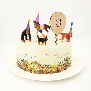 Könnte beinhalten: Ein weißer Kuchen mit bunten Streuseln am unteren Rand. Der Kuchen ist mit einer Holznummer 3 Ballon, einem Affen, einem Pferd, einem Hund und drei Partyhüten verziert.