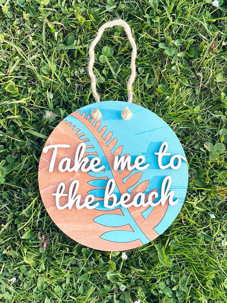 May include: Round wooden sign with the text "Take me to the beach" in white script. The sign features a blue and brown design with a fern-like shape. A jute rope is attached for hanging.