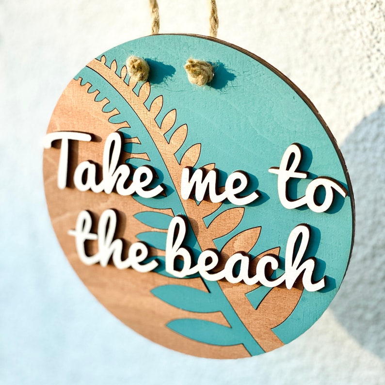 May include: Round wooden sign with the phrase "Take me to the beach" in white script. The sign features a teal and wood-toned design with a fern-like pattern and a jute rope for hanging.