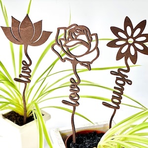 Könnte beinhalten: Drei Holzpflanzenpfähle mit Laser-Schnitzereien. Die Pfähle zeigen eine Lotusblume, eine Rose und eine Gänseblümchen. Die Worte "leben", "lieben" und "lachen" sind auf den Pfählen geschrieben.