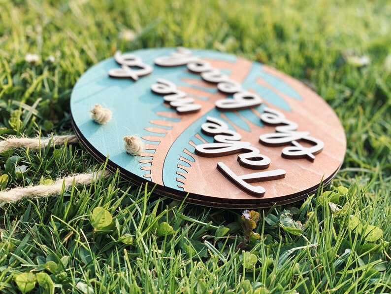 May include: Round wooden sign with the text "Take me to the beach" in a cursive font. The sign has a light blue center with a palm tree design and a natural wood border. Two small rope accents are attached.