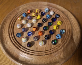 Puede incluir: Un tablero de juego de madera con un área de juego circular llena de canicas de colores. Las canicas son de varios tonos de azul, verde, rojo, amarillo, naranja y blanco. Algunas canicas tienen patrones o remolinos.