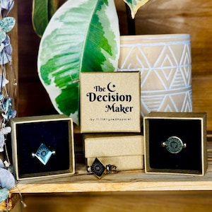 May include: Three small boxes with brown lids and a gold ring inside each. The boxes are stacked on a wooden shelf. The top box has the text "The Decision Maker - by 11:11AlignedApparel -" printed on the front.