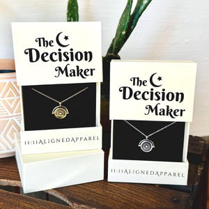 May include: Two white boxes with black text that reads "The Decision Maker" and a crescent moon symbol. Each box contains a silver necklace with a spinning charm that has a crescent moon symbol. The boxes are on a wooden surface.