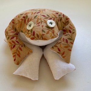 May include: A handmade frog plush toy with a tan fabric body featuring a fall leaf pattern in shades of red, orange, and green. The frog has white fabric legs and belly, and two large button eyes. A whimsical, decorative item.