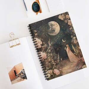 Puede incluir: Cuaderno de espiral con una ilustración celestial de una luna llena, estrellas y una figura con un vestido. La portada presenta elementos florales en tonos rosa y crema. El cuaderno está abierto, mostrando páginas en blanco.