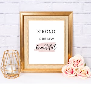 Op de afbeelding: Een ingelijste print met de woorden "STRONG IS THE NEW beautiful" in zwart script en een roze aquarel accent. De gouden lijst staat op een wit oppervlak met een gouden geometrische kandelaar en roze rozen.