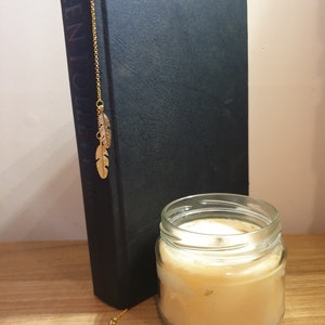 May include: A dark book with a gold feather bookmark. A lit candle in a glass jar sits beside the book on a wooden surface. The candle is a light yellow colour. The book has gold lettering.
