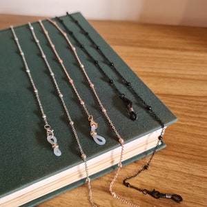 May include: Five different eyeglass chains with beaded details. The chains are silver, rose gold, gold, black, and a darker silver. The chains are laid out on a green book with the ends of the chains hanging off the edge of the book.