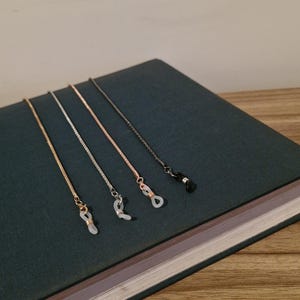 May include: Four eyeglass chains in gold, silver, rose gold, and black. Each chain has a small loop to attach to glasses. The chains are displayed on a dark green book.