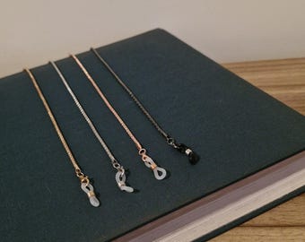 May include: Four eyeglass chains in gold, silver, rose gold, and black. Each chain has a small loop to attach to glasses. The chains are displayed on a dark green book.