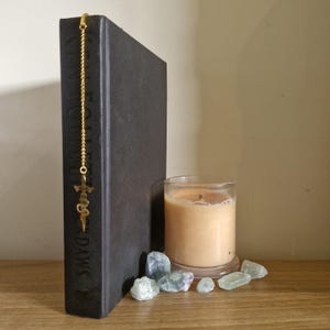 May include: A black hardback book with a gold bookmark featuring a sword design. The book is titled "The Cruel Prince" and is sitting on a wooden surface with a candle and green crystals.