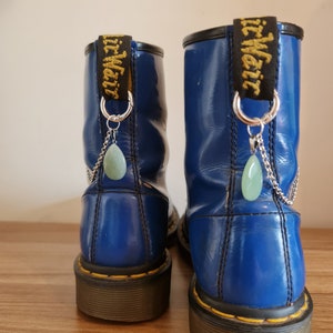May include: Two blue patent leather boots with black trim. Each boot features a silver chain and a pale green teardrop charm suspended from the pull loop. The pull loops are labelled "Dr. Martens" in yellow.