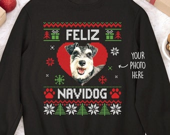 May include: A black long-sleeve sweatshirt featuring a festive holiday-themed graphic. The design includes a central red heart containing a dog portrait, the text "FELIZ NAVIDOG", and the text "YOUR PHOTO HERE" with an arrow pointing to the heart.