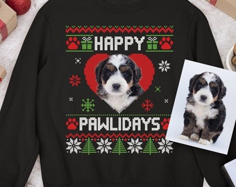 May include: A black long-sleeve sweatshirt featuring a knit-style graphic of a puppy inside a red heart. The text reads HAPPY PAWLIDAYS, surrounded by patterns of red paw prints, green gift boxes, white snowflakes, and green Christmas trees.